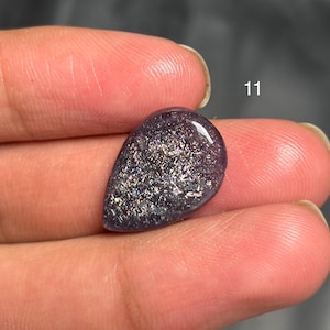 May include: A teardrop-shaped, polished gemstone with a deep purple hue and shimmering, silver-colored inclusions. The stone is held between fingers, with the number "11" visible in the background.