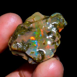 May include: A rough, natural opal with a green matrix. The opal displays a rainbow of colours, including red, orange, yellow, green, blue, and purple.