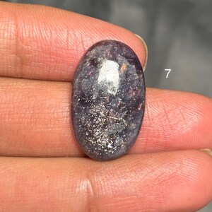 May include: An oval-shaped gemstone with a dark purple base and shimmering silver inclusions. The stone has a polished surface and is held between fingers. The number 7 is visible in the background.