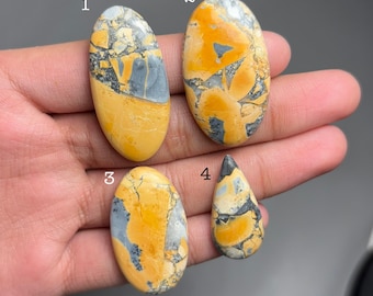May include: Four polished gemstones in shades of yellow, gray, and white. The stones are oval and teardrop-shaped, with unique patterns and textures. The gemstones are held in a hand, showcasing their natural beauty and variations. These stones are ideal for jewelry making.