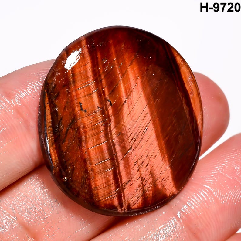 May include: A polished, oval gemstone in reddish-brown with a chatoyant effect. Parallel bands of colour create a striped look. The text "H-9720" is visible. The stone's surface reflects light, enhancing its natural patterns.