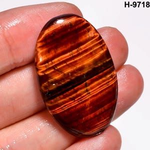 May include: An oval gemstone with a polished surface. The stone displays a pattern of parallel bands in shades of red, orange, and black. The text "H-9718" is visible in the upper right corner. The gemstone is held in a hand.