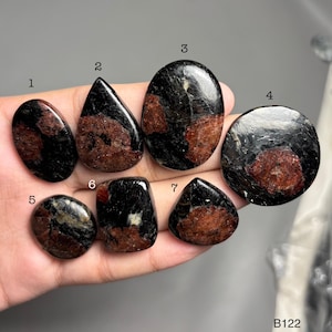 Puede incluir: Colección de siete piedras preciosas pulidas de varias formas, incluyendo ovaladas, en forma de lágrima y redondas. Las piedras son principalmente negras con inclusiones de color marrón rojizo, mostrando patrones naturales. Las piedras preciosas se muestran en una mano.