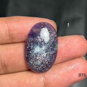 May include: An oval-shaped gemstone with a shimmering, speckled appearance. The stone features a blend of purple, blue, and silver hues, with a polished, reflective surface. The number "1" and the letters "B73" are visible.