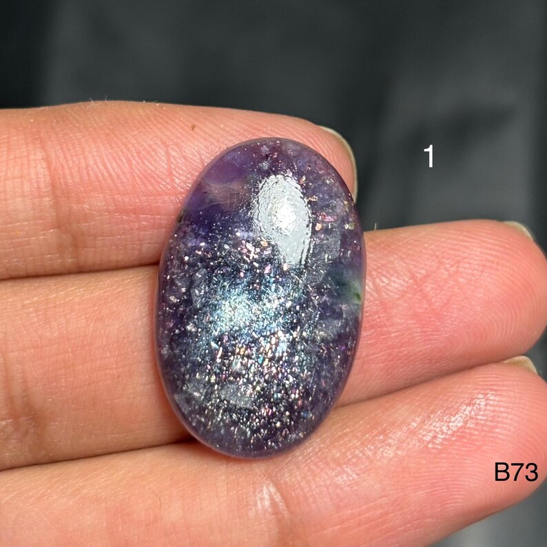 May include: An oval-shaped gemstone with a shimmering, speckled appearance. The stone features a blend of purple, blue, and silver hues, with a polished, reflective surface. The number "1" and the letters "B73" are visible.