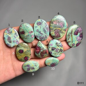 May include: A collection of ten polished ruby zoisite gemstones in various shapes, including oval and teardrop. The stones feature a mix of green and pink hues, with the ruby inclusions creating a striking contrast. The gemstones are displayed on a hand.