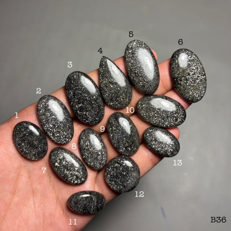 May include: A collection of polished black gemstones with shimmering gold flecks, held in a hand. The stones vary in shape, including ovals, teardrops, and a round stone. The gemstones are numbered for identification.
