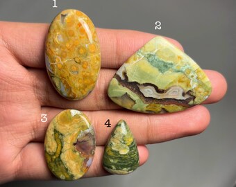 May include: Four polished gemstones in various shapes and colors. The stones include oval, triangular, and teardrop shapes, with colors ranging from yellow and green to brown and white. The gemstones are displayed against a neutral background.