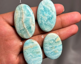 May include: Four oval gemstones in shades of blue and white are shown. The polished stones have unique patterns and textures. The gemstones are held in a hand, showing their size and shape. The stones are likely for jewellery.