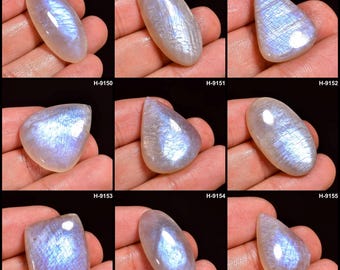 May include: Nine polished moonstone cabochons in various shapes, including oval, teardrop, and square. Each stone displays a milky white base with iridescent blue and silver flashes. The stones are held in a hand against a black background.