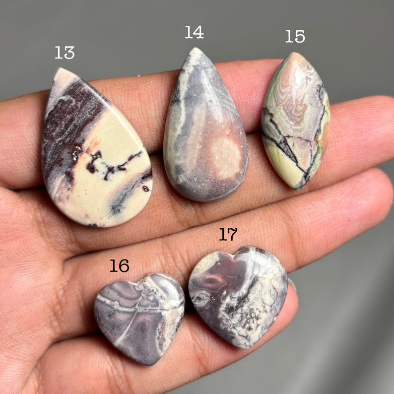 May include: A collection of polished gemstones in various shapes. The stones include teardrop, oval, and heart shapes. Colors range from cream and beige to gray and purple, with unique veining patterns. The gemstones are numbered 13, 14, 15, 16, and 17.