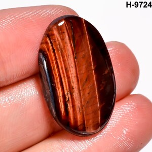 May include: Oval-shaped red tiger's eye gemstone with dark brown and black stripes. The polished stone has a glossy finish and is held in a hand. The text "H-9724" is in the upper right corner.