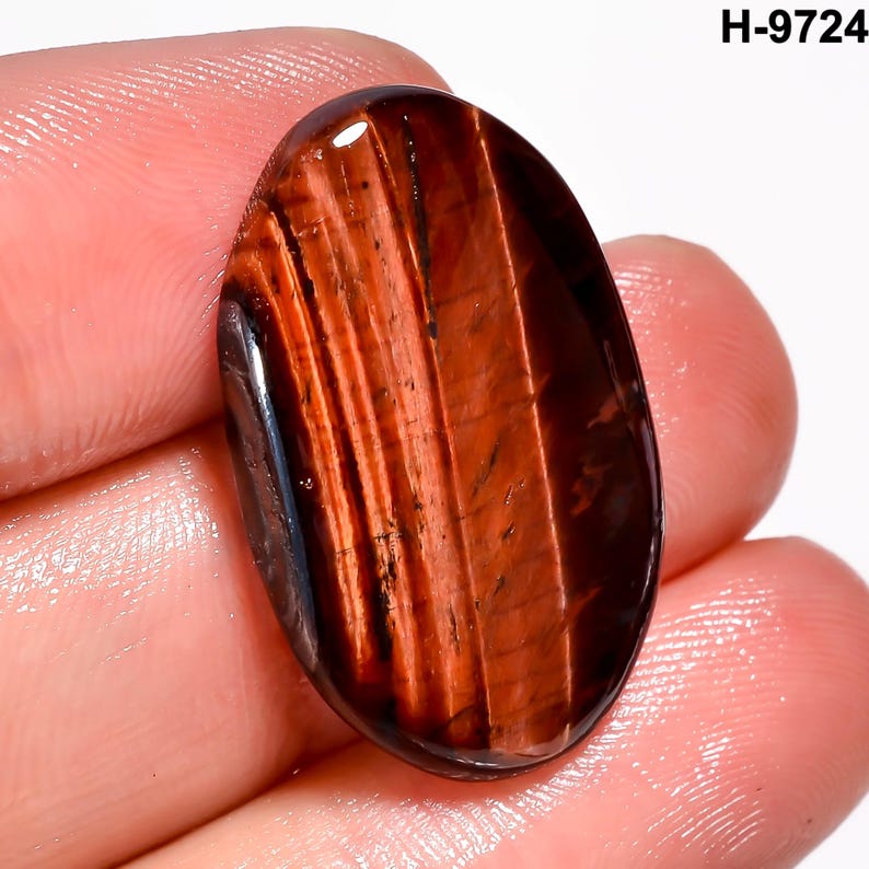 May include: Oval-shaped red tiger's eye gemstone with dark brown and black stripes. The polished stone has a glossy finish and is held in a hand. The text "H-9724" is in the upper right corner.
