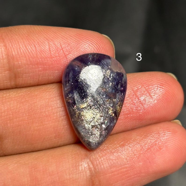 May include: A teardrop-shaped gemstone with a deep purple hue, accented by shimmering gold and silver inclusions. The stone is held in a hand, with the number "3" visible in the background. The polished surface reflects light, highlighting the unique patterns.