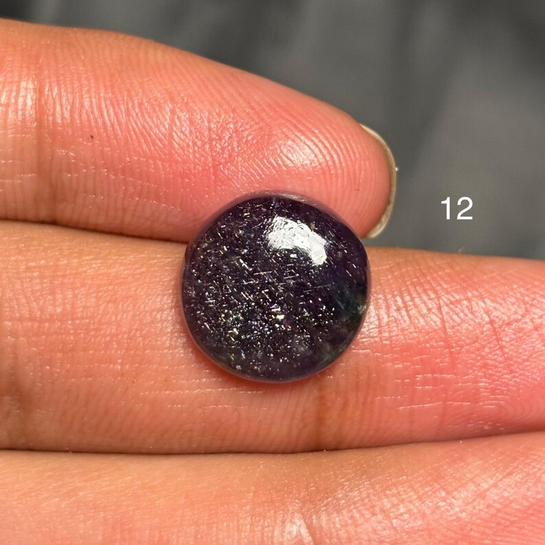 May include: A round, polished gemstone with a deep purple hue, speckled with shimmering silver and iridescent flecks. The gem is held in a hand, with the number "12" visible in the background. The stone has a smooth, domed surface.