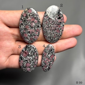 May include: Four oval gemstones with a speckled pattern of pink and black. The stones are held in a hand against a grey background. The gemstones vary in size and shape, with a polished surface. The bottom right corner of the image has the text "B 99".