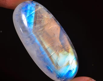 Könnte beinhalten: Ein glatter, ovaler Mondstein-Cabochon mit einer milchig weißen Basis und schillernden blauen und goldenen Blitzen.