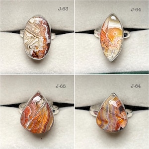 Puede incluir: Cuatro anillos de plata con piedras preciosas de ágata bandeada en formas ovalada, marquesa y de lágrima. Las piedras muestran patrones de remolinos en blanco, naranja y marrón. Cada anillo tiene el texto J-63, J-64 o J-65.