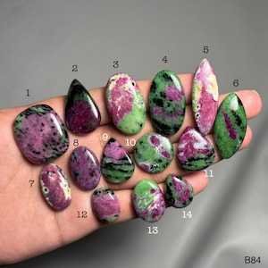 May include: A selection of polished ruby zoisite gemstones in diverse shapes, such as oval, teardrop, and irregular forms. The stones exhibit a blend of pink, green, and black hues, with visible patterns and textures. The gemstones are held in a hand.