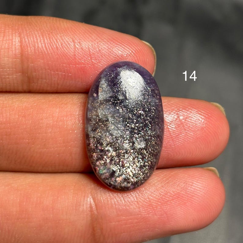 May include: An oval-shaped gemstone with a dark purple hue, speckled with silver and iridescent glitter. The gemstone is held between fingers, with the number "14" visible in white.