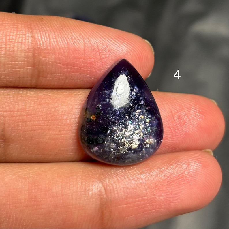 May include: A teardrop-shaped gemstone with a deep purple hue, speckled with silver and gold flecks. The stone has a polished, reflective surface, and the number "4" is visible in the upper right corner.