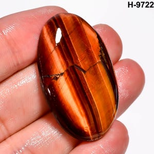 May include: An oval tiger's eye gemstone with a polished surface. The stone features bands of reddish-brown and gold colours. The gemstone is held in a hand, and the text "H-9722" is visible in the upper right corner.
