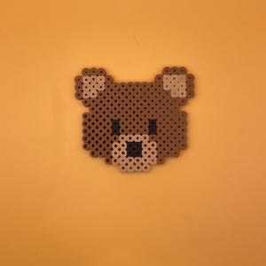 Puede incluir: Una cara de oso marrón hecha con cuentas perler marrones, beige y negras.