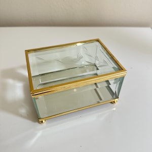 May include: A rectangular glass jewelry box featuring a gold metal frame and four small gold ball feet. The hinged lid is made of clear glass with a delicate etched design of a bird perched on a branch, providing a clear view of the interior.