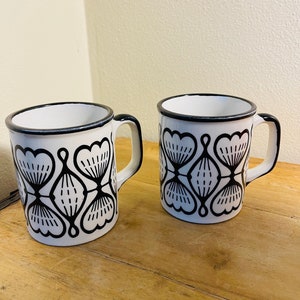 May include: Two white ceramic mugs with black geometric designs. The mugs have black rims and handles.