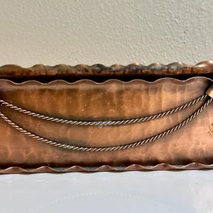 May include: A copper-toned metal planter with a scalloped rim and decorative rope and diamond accents. The planter has scrolled handles on each end. The hammered metal surface gives the planter a rustic appearance. The planter is approximately 18 inches long.