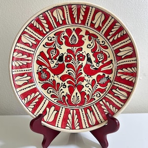 May include: A decorative ceramic plate with a hand-painted design. The plate features a cream-colored center with red floral and bird motifs. The border is red with white leaf-like patterns. The plate is displayed on a dark red stand.