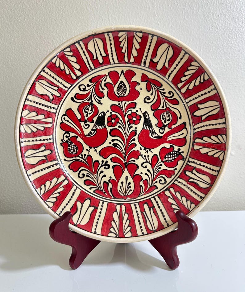 May include: A decorative ceramic plate with a hand-painted design. The plate features a cream-colored center with red floral and bird motifs. The border is red with white leaf-like patterns. The plate is displayed on a dark red stand.