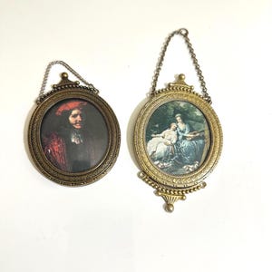 May include: Two oval wall frames with metallic gold-toned finishes and hanging chains. The left frame contains a portrait of a person in a red hat and dark clothing. The right frame features a scene of two figures in a garden setting.