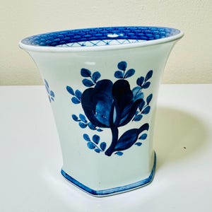 May include: White ceramic vase with a blue floral design. The vase has a flared shape with a blue patterned rim and a blue floral motif on the front. The vase is approximately 8 inches tall.