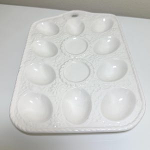 May include: A white ceramic egg holder with a floral pattern. It features 12 egg-shaped recesses and two circular spaces. The tray has a handle at the top and a decorative edge.