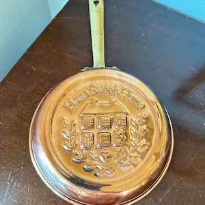 May include: A round copper pan with a brass handle featuring an embossed design of a house and the text Home Sweet Home. The center of the pan displays a detailed house illustration surrounded by leafy branches and a small heart shape at the base.
