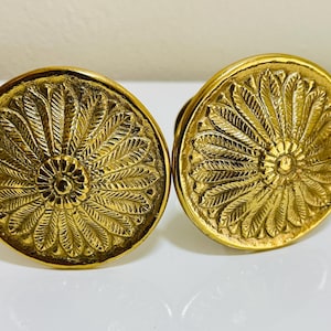 May include: Two antique gold-colored cabinet knobs. Each knob is round with a detailed, raised floral design. The knobs are made of metal and have a shiny finish. The knobs are likely for a drawer or cabinet.