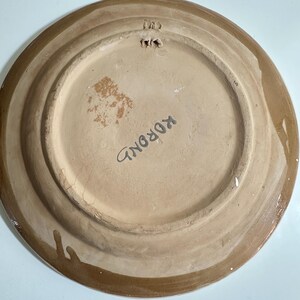 May include: A ceramic plate with a light brown center and a darker brown rim. The word "KOROND" is written in black on the plate's surface. The plate has a hole for hanging.