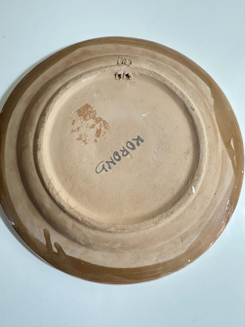 May include: A ceramic plate with a light brown center and a darker brown rim. The word "KOROND" is written in black on the plate's surface. The plate has a hole for hanging.