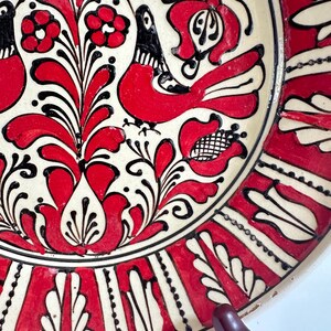 May include: A decorative ceramic plate with a cream-colored base and a vibrant red and black floral design. The plate features stylized birds and flowers, with a black border and red accents. The design is hand-painted, showcasing a folk art style.