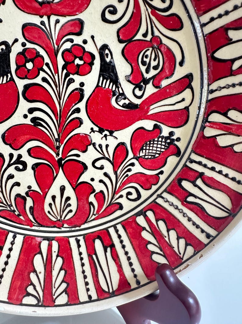 May include: A decorative ceramic plate with a cream-colored base and a vibrant red and black floral design. The plate features stylized birds and flowers, with a black border and red accents. The design is hand-painted, showcasing a folk art style.