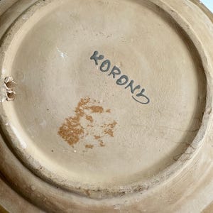 May include: The underside of a ceramic plate with a light brown base and a darker brown rim. The word "KORON" is written in black script. The plate has a circular shape with a raised edge.
