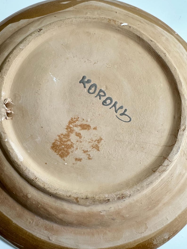 May include: The underside of a ceramic plate with a light brown base and a darker brown rim. The word "KORON" is written in black script. The plate has a circular shape with a raised edge.