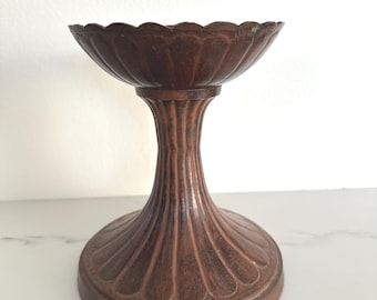 May include: A decorative, antique-style pedestal bowl in a warm, reddish-brown hue. The bowl has a scalloped rim and a fluted stem, resting on a rounded base with a similar fluted design. The metal appears aged, adding to its vintage charm.