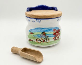 May include: A white ceramic salt cellar with a blue rim and a wooden lid. The front features a hand-painted scene of a person on a bicycle with red trees and green grass. The text "île de Ré" is written in blue script. A small wooden scoop is included.