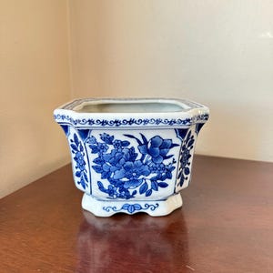 May include: A hexagonal ceramic planter featuring a white base with intricate blue floral patterns and decorative border detailing. The vessel has a flared rim and a raised base, showcasing detailed botanical illustrations on its exterior.