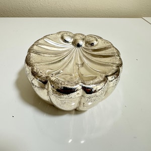 May include: A round, lidded glass container with a metallic silver finish and a distressed, antique-style texture. The lid features a raised, scalloped shell-like design radiating from a central point, creating a decorative, textured surface.