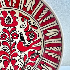 May include: A decorative ceramic plate with a vibrant red, black, and white floral design. The plate features a central motif of stylized flowers and a bird, surrounded by a red border with white leaf-like patterns. The rim is a creamy white.