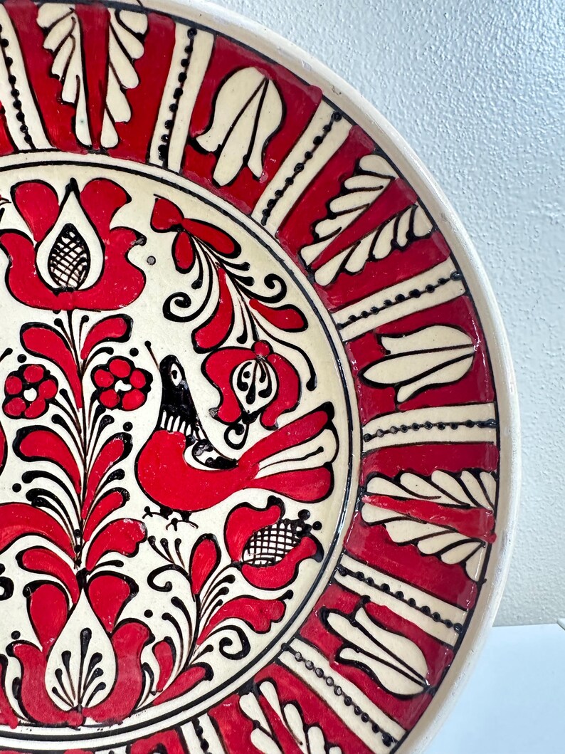 May include: A decorative ceramic plate with a vibrant red, black, and white floral design. The plate features a central motif of stylized flowers and a bird, surrounded by a red border with white leaf-like patterns. The rim is a creamy white.