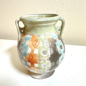 May include: A ceramic vase with a bulbous body and two handles. The vase features a patchwork design in shades of orange, green, blue, and brown, with white and gray accents. The rim and handles are a light green color.
